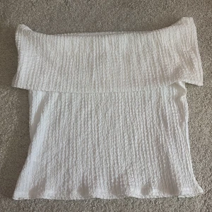 Vit offshoulder topp från H&M - Säljer en vit offshoulder topp från H&M i storlek L. Toppen har en snygg ribbad struktur och är axelbandslös med bred volangkant. Materialet är mjukt och lätt.