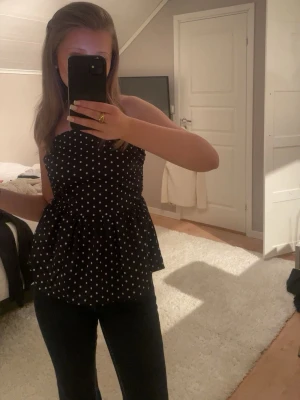 Svart prickig bandeau topp - Säljer en svart bandeau topp med vita prickar och volang nertill. Toppen är axelbandslös och har en söt, lite utställd form som ger en feminin siluett. Perfekt att styla med jeans eller kjol för en trendig look.