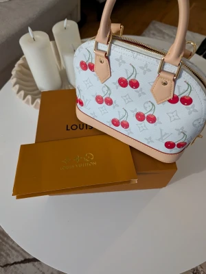 Väska  - Supersöt vit handväsk med körsbärsmönster och beige detaljer. Väskan har en stor rosett framtill, guldfärgade metalldetaljer och röd insida. Perfekt storlek för det viktigaste och har både handtag och axelrem.