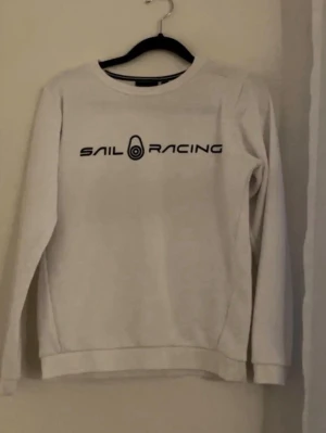 Vit Sail Racing sweatshirt - Vit sweatshirt från Sail Racing med svart logotyptryck på bröstet. Tröjan har rund halsringning och långa ärmar. Tillverkad i mjuk bomull för en skön och avslappnad stil. Perfekt för dig som gillar sportiga och stilrena plagg. Skicket är ungefär 9,5/10 skriv gärna för intresse elr pris diskutering