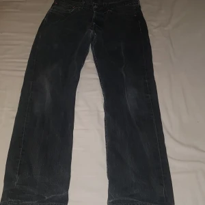 Levi's 501 svarta jeans W31 L30 - Klassiska svarta Levi's 501 jeans med rak passform och knappgylf. Jeansen har fem fickor, ikonisk röd Levi's-tag på bakfickan och patch bak i midjan. Tillverkade i slitstarkt bomullsjeansmaterial. Perfekta för en avslappnad och tidlös look.