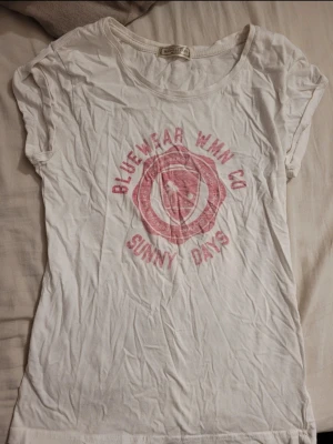 Vit t-shirt Bluewear WMN Co - Vit t-shirt från Bluewear WMN Co med tryck i rosa och texten 'Sunny Days' framtill. Klassisk rund halsringning och korta ärmar. Perfekt för en avslappnad look och enkel att matcha till jeans eller shorts.