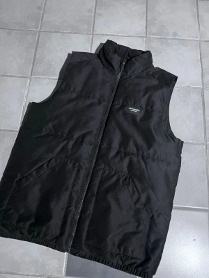 Svart dunväst Abercrombie & Fitch - Säljer en svart dunväst från Abercrombie & Fitch med hög krage och dragkedja framtill. Västen har två stora fickor och en stilren look, perfekt att slänga över en hoodie eller tröja. Klassisk och enkel design som funkar till det mesta.