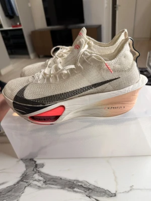 Nike Alphafly 3 löparskor vit/svart - Nike Alphafly 3 löparskor i vitt med svart swoosh och detaljer i neonrosa. Ovandelen är stickad och ventilerande, med snörning och hög, vadderad häl. Mellansulan är tjock och har synlig Zoom Air-enhet för extra respons. Perfekta för dig som vill maxa löpningen.