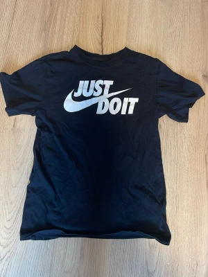 Svart Nike t-shirt Just Do It S - Säljer en svart t-shirt från Nike i storlek S med det klassiska 'Just Do It'-trycket i vitt på bröstet. T-shirten har rund hals och korta ärmar, perfekt för en sportig och avslappnad stil.