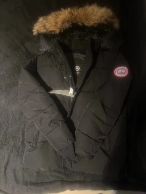 Svart dunjacka från Canada Goose M - Säljer en svart dunjacka från Canada Goose med ikonisk patch på ärmen och stor huva med brun fuskpäls. Jackan har dragkedja, långa ärmar och är riktigt tjock och varm. Perfekt för kalla vinterdagar och har en klassisk, clean look.