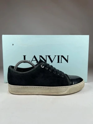 Svarta sneakers från Lanvin(DBB1) - Tjena säljer ett par äkta Lanvin sneakers i svart mocka med detaljer i blankt skinn på tån och snörning framtill. Sulan är vit vilket ger en clean och stilren look. Perfekta för dig som gillar exklusiva och snygga sneakers med lyxig känsla. Kommer med original box.