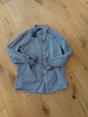 Randig skjorta från Tommy Hilfiger - Snygg skjorta från Tommy Hilfiger med svartvita ränder och klassisk krage. Skjortan har långa ärmar, knappar framtill och är tillverkad i mjuk bomull. Perfekt för dig som gillar stilrena och tidlösa plagg. Helt ny, lappen sittter fof på! Pris kan diskuteras!