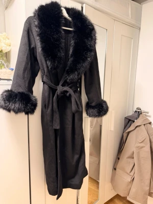 Svart kappa med f pälsdetaljer - Säljer en svart lång kappa med bred krage och ärmslut i fluffig fuskpäls. Kappan har knytband i midjan för snygg passform och är helsvart med en elegant look. Perfekt för dig som vill ha en statement jacka till höst och vinter.