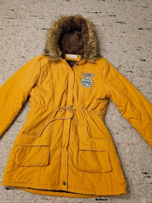 Gul parkasjacka med fuskpäls - Gul parkasjacka med bruna fuskpälsdetaljer i huvan och mjukt teddyfoder. Jackan har snörning i midjan, stora fickor framtill, dragkedja och knappar. Dekorativa patches på bröstet ger en cool army-vibe. Perfekt för kalla dagar.
