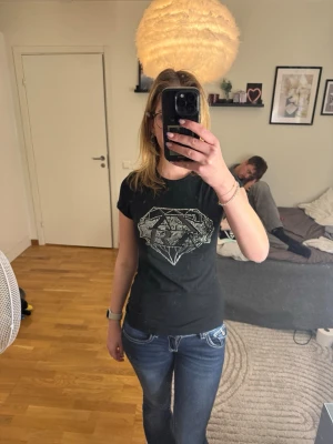 Svart t-shirt med diamanttryck Zara - Svart t-shirt från Zara Evening Collection med ett stort diamanttryck framtill i silverfärgade nitar. Modellen är kortärmad och har rund hals. Materialet är mjuk bomull som sitter skönt mot kroppen. Perfekt för dig som gillar lite extra bling.