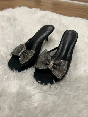 Svarta pumps med glittrig rosett - Säljer ett par svarta pumps med öppen tå och låg klack. Skorna har en stor glittrig rosett framtill som ger en extra touch. Materialet är troligtvis syntet med meshdetaljer runt rosetten. Perfekta för dig som vill sticka ut lite extra.