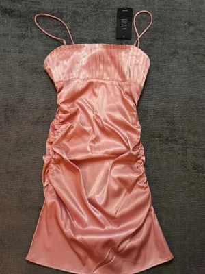 Rosa satin klänning  - Supersnygg rosa kortklänning från NAKD i glansigt satinmaterial. Klänningen har tunna axelband, plisserad överdel och rynkade sidor som ger en cool och trendig look. Perfekt för dig som vill sticka ut lite extra!