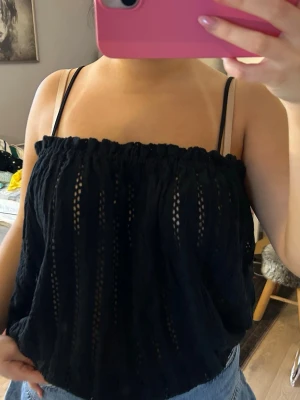 Svart ärmlös linnetopp med hålmönster - Säljer en svart linnetopp med tunna axelband och elastisk kant upptill. Toppen har ett snyggt hålmönster i vertikala ränder och är luftig och perfekt för varma dagar. Passar till jeans eller kjol för en chill look.