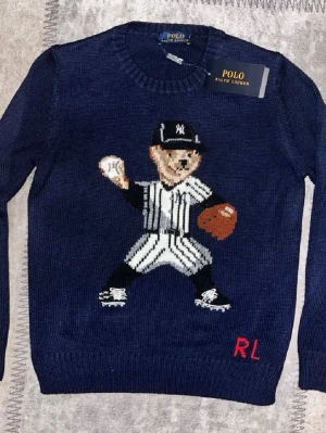 Mörkblå stickad tröja Polo Ralph Lauren - Säljer en mörkblå stickad tröja från Polo Ralph Lauren med en broderad björn i basebollkläder på framsidan. Tröjan har rund halsringning, långa ärmar och RL-broderi i rött nedtill. Perfekt för dig som gillar sportig och lekfull stil.