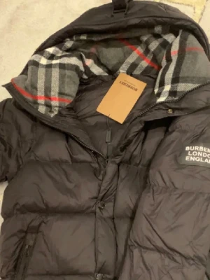 Svart pufferjacka från Burberry - Säljer en svart pufferjacka från Burberry i storlek S. Jackan har klassiskt rutigt foder i grått, vitt och rött, dragkedja och knappar framtill samt två sidofickor med dragkedja. Perfekt för kalla dagar och riktigt snygg med den ikoniska Burberry-looken. För fler bilder och funderingar skrev gärna