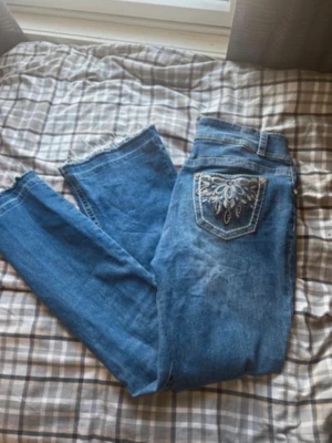 Blå jeans från Copper Flash med broderi - Säljer ett par blå jeans från Copper Flash med snyggt broderade bakfickor i vitt och silver. Jeansen har normal passform, dragkedja och råa kanter vid bensluten för en trendig look. Perfekta för dig som gillar detaljer och vill sticka ut lite extra. Dessa är köpta i usa