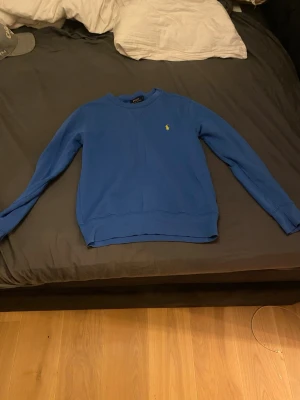 Blå sweatshirt från Polo Ralph Lauren - Klassisk blå sweatshirt från Polo Ralph Lauren i storlek S. Tröjan har rund halsringning, långa ärmar och den ikoniska gula logon broderad på bröstet. Tillverkad i mjuk bomull för en skön och avslappnad look.