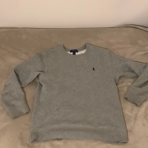 Grå sweatshirt från Polo Ralph Lauren - !!!ÄKTA!!!Grå sweatshirt från Polo Ralph Lauren med klassisk rund hals och långärmad passform. Tröjan har en broderad marinblå Polo-logga på bröstet och är tillverkad i mjuk bomullsmix. Perfekt för chill dagar och enkel att matcha med jeans eller joggers.