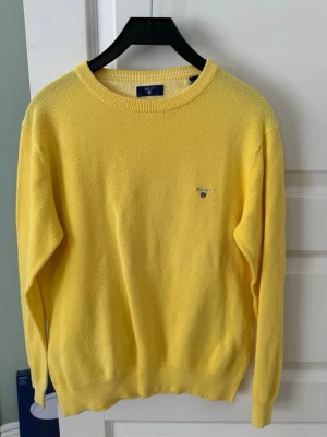 Gul tröja från GANT - Säljer en ljusgul stickad tröja från GANT med rund halsringning och diskret broderad logga på bröstet. Tröjan har lång ärm och ribbade muddar vid ärmslut och nederkant. Perfekt för dig som vill ha en färgklick i garderoben.
