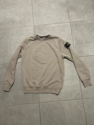 Beige sweatshirt från Stone Island - Säljer en beige sweatshirt från Stone Island med det klassiska logotypmärket på ärmen och ett stort broderat emblem på bröstet. Tröjan har rund hals, ribbade muddar och är långärmad. Perfekt för dig som gillar stilrena och exklusiva plagg.