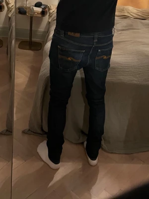 Mörkblå skinny jeans från Nudie - Säljer ett par mörkblå lean Dean jeans från Nudie med klassiska kontrastsömmar och deras ikoniska broderi på bakfickorna. Storleken är 30W/34L men sitter mer som 32 i längden. Personen på bilden är 185 lång och väger 72 kg. Tveka inte med att ställa frågor.