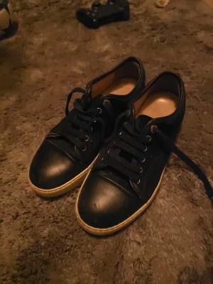 Svarta Lanvins  - Säljer ett par svarta Lanvins med vit sula och rund tå. Skorna har snörning med svarta skosnören och metallöljetter. Ovandelen är i skinn och textil, vilket ger en clean och klassisk look. Passar perfekt till jeans eller shorts. Skriv vid frågor o liknande. Skon har en pytte defekt längst fram vilket inte påverkar använingen!