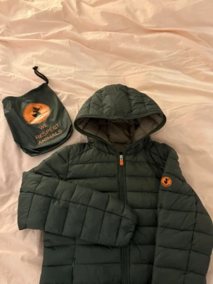 Mörkgrön pufferjacka från Save The Duck - Säljer en mörkgrön pufferjacka från Save The Duck med huva och orange logga på ärmen. Jackan är quiltad, har dragkedja framtill och är tillverkad i syntetmaterial med Plumtech-fyllning. Medföljer praktisk förvaringspåse. Perfekt för kyliga dagar. 