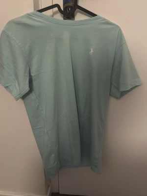 Ljusblå t-shirt från Polo Ralph Lauren - Snygg ljusblå t-shirt från Polo Ralph Lauren med klassisk vit broderad logga på bröstet. T-shirten har rund halsringning och korta ärmar. Tillverkad i mjuk bomull som känns skön mot huden. Perfekt för en clean och stilren look.