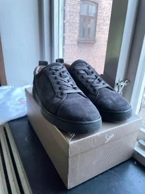 Mörkgrå mocka sneakers från Louboutin - Snygga mörkgrå sneakers från Christian Louboutin i mjuk mocka med klassisk röd sula. Skorna har rund tå, snörning och diskret design som passar till många stilar. Kommer med originalkartong och dustbag.