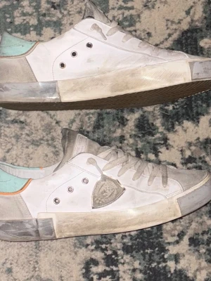 Philippe Model vita sneakers med mintdetalj - Säljer ett par vita Philippe Model sneakers med grå mockadetaljer och mintgrön häl. Skorna har snörning, rund tå och en platt sula. Sidan pryds av den klassiska sköldloggan och orange detalj vid hälen. Perfekta för dig som gillar stilrena men unika sneakers.