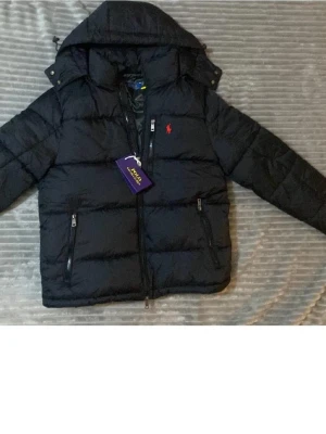 Svart pufferjacka från Polo Ralph Lauren - Säljer en svart pufferjacka från Polo Ralph Lauren med huva och dragkedja. Jackan har två framfickor med dragkedja och den klassiska röda loggan på bröstet. Perfekt för kalla dagar och riktigt snygg till alla outfits.