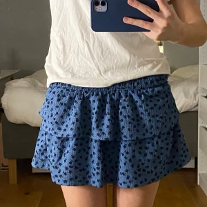 Blå volangkjol från Zara  - Supersöt blå kortkjol med volanger och små mörka blommor från Zara. Kjolen har resår i midjan för skön passform och är gjord i ett mjukt, strukturerat tyg. Perfekt för dig som gillar en lekfull och trendig stil.