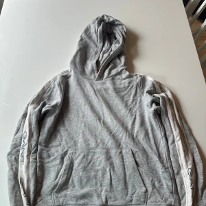 Grå hoodie från GANT med logga - Snygg grå hoodie från GANT med huva och känguruficka. Ärmarna har vita ränder med GANT-logga längs sidorna. Använd ett fåtal gånger så i väldigt fint skick. 