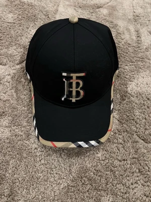 Burberry keps - Säljer en snygg Burberry keps med svart ovandel och klassiskt beige, röd och svart rutigt mönster pá skärmen. Framtill finns ett broderat TB-monogram i beige och baktill stär Burberry broderat. Justerbar rem med metallspänne och rutigt tyg.