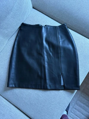 Svart kort kjol i skinnimitation BikBok - Säljer en svart kort kjol från BikBok i skinnimitation. Kjolen har en clean look med en liten slits framtill som ger en edgy vibe. Perfekt att styla med chunky boots eller en oversized tröja.