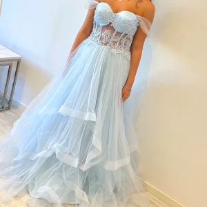 Ljusblå långklänning med spetsdetaljer, med korsett där bak - Drömmig ljusblå långklänning med voluminös tyllkjol och axelbandslös överdel. Livet har vackra spetsapplikationer och transparenta partier som ger en modern och romantisk känsla. Perfekt för bal eller speciella tillfällen.