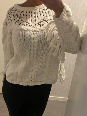 Vit stickad tröja med fransar - Säljer en vit stickad tröja med snygga virkade detaljer upptill och flätstickade partier. Tröjan har långa ärmar och coola fransar längs sidorna och ärmarna för en unik look. Perfekt till jeans eller kjol för en trendig outfit.