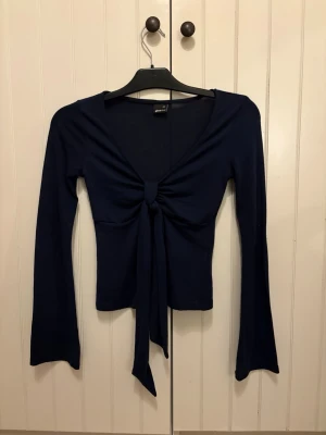 Mörkblå knytblus från Gina Tricot - Säljer en mörkblå långärmad blus från Gina Tricot med snygg knytning framtill och v-ringad hals. Toppens material är mjukt och stretchigt, vilket gör den bekväm att bära. Perfekt till jeans eller kjol för en trendig look.