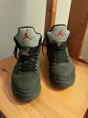 Nike Air Jordan 5 Retro Olive strl.38,5 - Säljer ett par Nike Air Jordan 5 Retro Olive med olivgröna mockapaneler, svarta snören och meshdetaljer på sidorna. Tungorna är grå med röd Jumpman-logga och sulan har genomskinliga partier samt orange detaljer. Klassisk high-top siluett och ikonisk design.