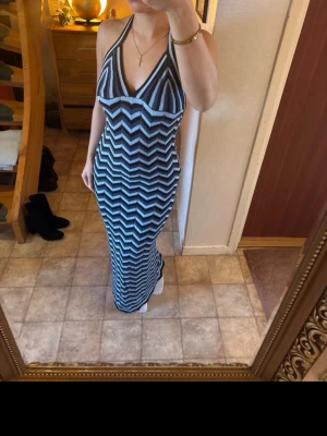 Randig långklänning med öppen rygg - Supersnygg långklänning med svart och vit zigzag-randigt mönster. Klänningen har halterneck, v-ringning och öppen rygg med knyt i nacken. Sitter tight och framhäver figuren. Perfekt för dig som vill sticka ut med en cool och trendig look.