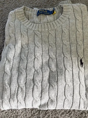 Grå kabelstickad tröja Polo Ralph Lauren - Säljer en klassisk grå kabelstickad tröja från Polo Ralph Lauren. Tröjan har rund halsringning, ribbade muddar och det ikoniska broderade logotypen på bröstet. Perfekt för lager på lager under höst och vinter.