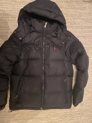 Svart pufferjacka från Polo Ralph Lauren - Säljer en marinblå pufferjacka från Polo Ralph Lauren med huva och dragkedja. Jackan har det klassiska röda Polo-logot broderat på bröstet och flera praktiska fickor med dragkedja. Perfekt för kalla dagar och riktigt snygg till Stockholm-stilen eller grish stilen