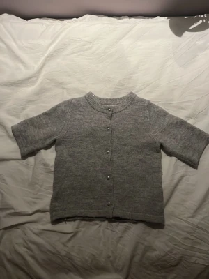 Grå kofta med kort ärm och knappar - Mysig grå kofta med korta ärmar och rund hals. Koftan har knappar framtill och är stickad i ett mjukt material som passar perfekt för lager på lager. Enkel och stilren design som funkar till det mesta.