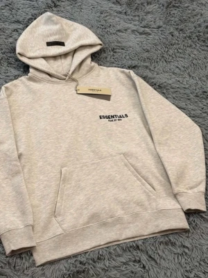 Essentials Fear of God Hoodie – Beige• Fear of God ESSENTIALS Hoodie i Ljusbeige• Essentials Hoodie med Bröstlogga – Beige• Fear of God Essentials Pullover Hoodie• Essentials FOG Hoodie – Klassisk Beige - Beige hoodie från Fear of God ESSENTIALS i högkvalitativ och mjuk bomullsblandning. Tröjan har en avslappnad, modern passform med ribbade muddar och nederkant för extra komfort. Klassisk känguruficka framtill samt huva utan snören för en clean look. Diskret ESSENTIALS-logotyp tryckt på bröstet ger ett stilrent och tidlöst uttryck. Perfekt för vardagsbruk och enkel att matcha med både jeans och mjukisbyxor.