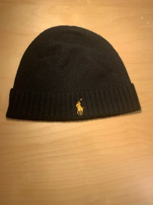 Svart mössa från Polo Ralph Lauren - Säljer en svart mössa från Polo Ralph Lauren i mjuk merinoull. Klassisk ribbad kant och ikoniska broderade loggan i gult framtill. Perfekt accessoar för kyliga dagar och enkel att matcha med olika stilar.