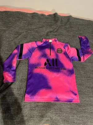 PSG Mbappé träningströja barnstorlek M - Säljer en PSG träningströja med Mbappé och nummer 7 på ryggen. Tröjan har ett färgstarkt rosa och lila mönster, svart PSG-logga och sponsortryck framtill samt halv dragkedja vid halsen. Tillverkad i mjuk polyester, perfekt för fotbollsträning. OBS är detta barnstorlek M! Hör av dig vid fler frågor! 