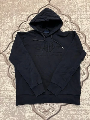 Mörkblå Gant Hoodie - Mörkblå hoodie från GANT med broderad logga och text på bröstet. Klassisk modell med huva och dragsko, ribbade muddar och mjukt material. Perfekt för en chill och stilren look. Priset är inte i hugget’ så kan komma överens om något! den har tappat lite färg så därför de ser ut så på bilden. Den är mörk blå 