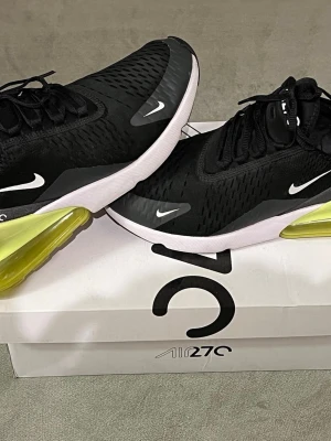Nike Air Max 270 svarta, gröna sneakers  - Nike Air Max 270 sneakers i svart mesh med vita detaljer och en stor, genomskinlig limegrön Air-enhet i hälen. Skorna har snörning, rund tå och vit sula. Perfekta för dig som vill ha en sportig och modern look med maximal komfort. Kommer med den original skokartong. 
