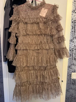 Beige spetsklänning från By Malina - Supersnygg beige spetsklänning från By Malina med volanger och hög hals. Klänningen har lång ärm, dragkedja bak och är täckt av vackra spetsdetaljer. Perfekt för dig som vill sticka ut med en romantisk och trendig look. Köpt för 2799 och aldrig använd med prislapp kvar 🩷